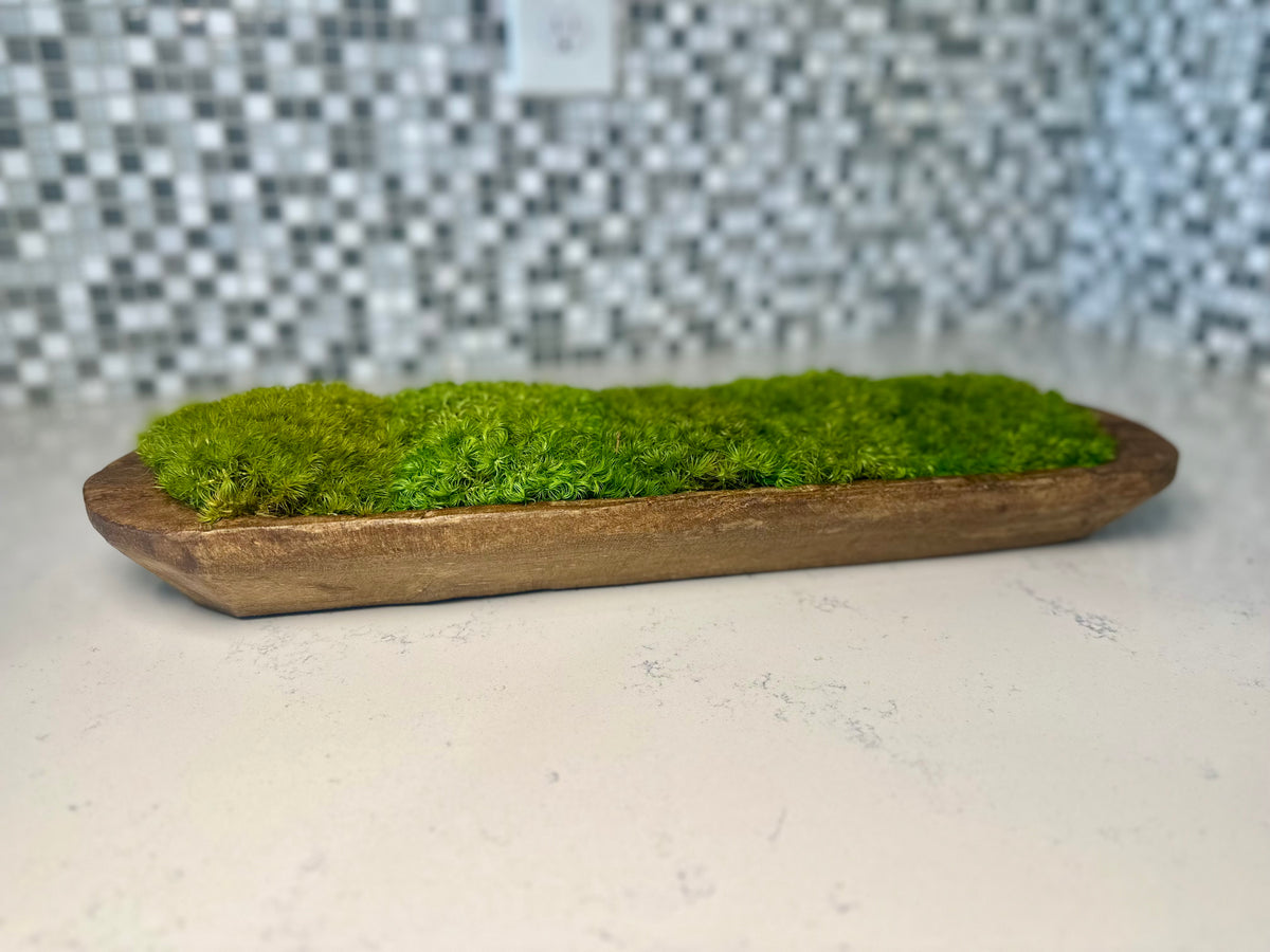 Side view of a rustic brown wood dough bowl with rounded green preserved moss mounds on a white countertop.