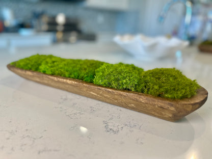 Angled view of a long hand-carved wood dough bowl filled with green preserved moss on a white countertop.