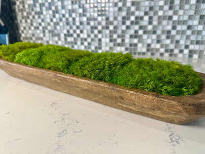 Close-up side view of green preserved moss mounds in a long rustic wood dough bowl on a white countertop.
