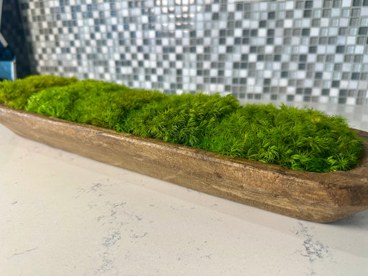 Close-up side view of green preserved moss mounds in a long rustic wood dough bowl on a white countertop.