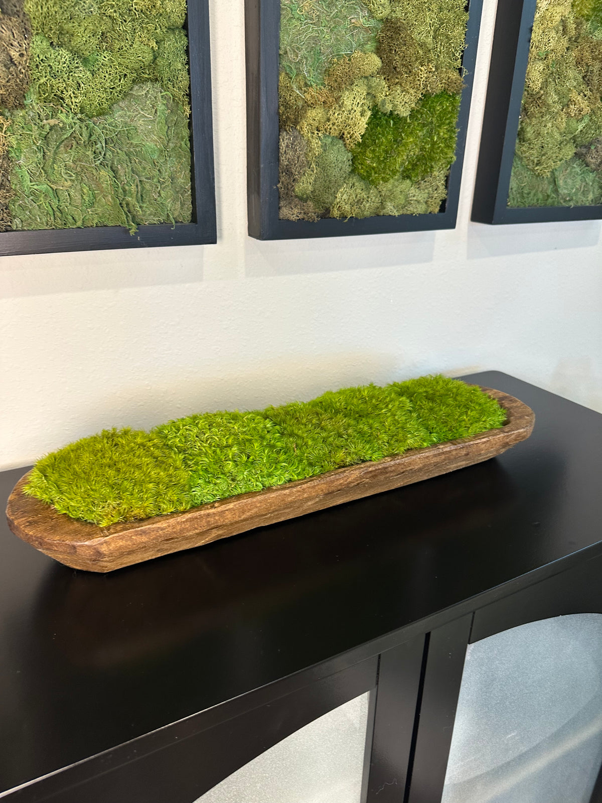 Rustic wood dough bowl filled with green preserved moss, displayed on a black console beneath framed moss art.