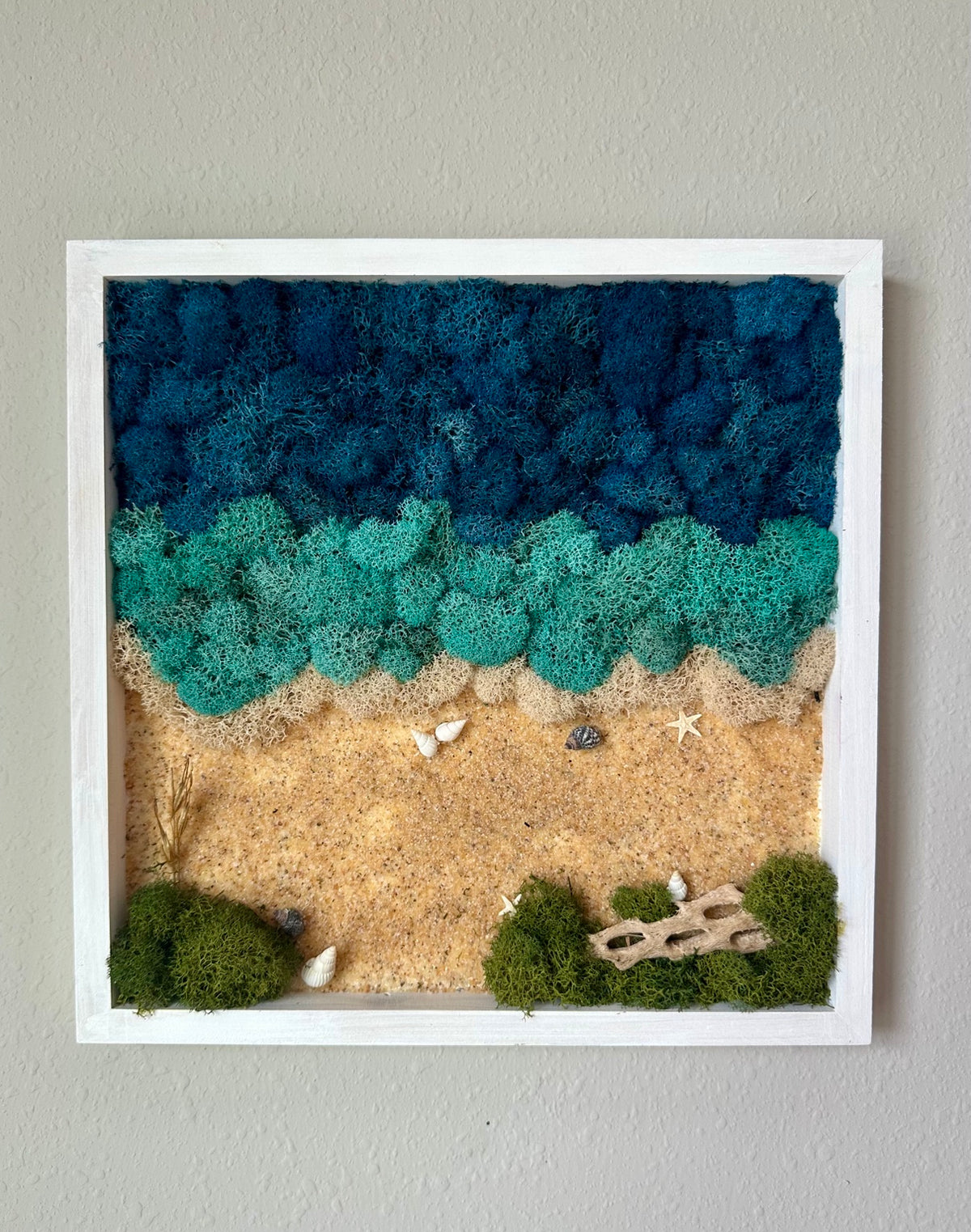 Square white-framed beach scene with deep blue and turquoise preserved moss waves, an ivory shoreline, sand, shells, green moss, and a driftwood accent.